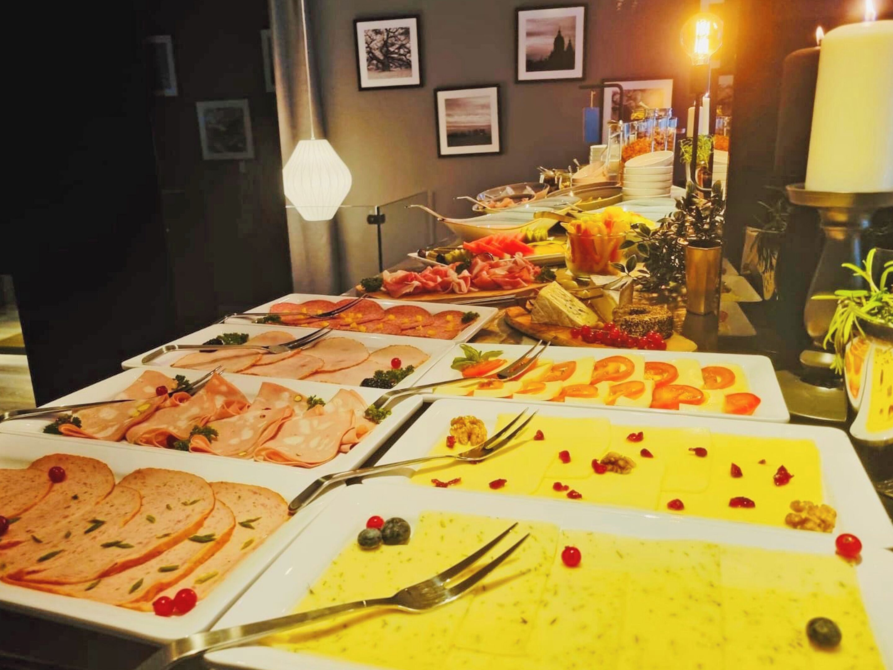 Een buffettafel met diverse etenswaren, waaronder vlees, kazen en fruit, in een gedimd verlichte ruimte met ingelijste foto’s aan de muur.