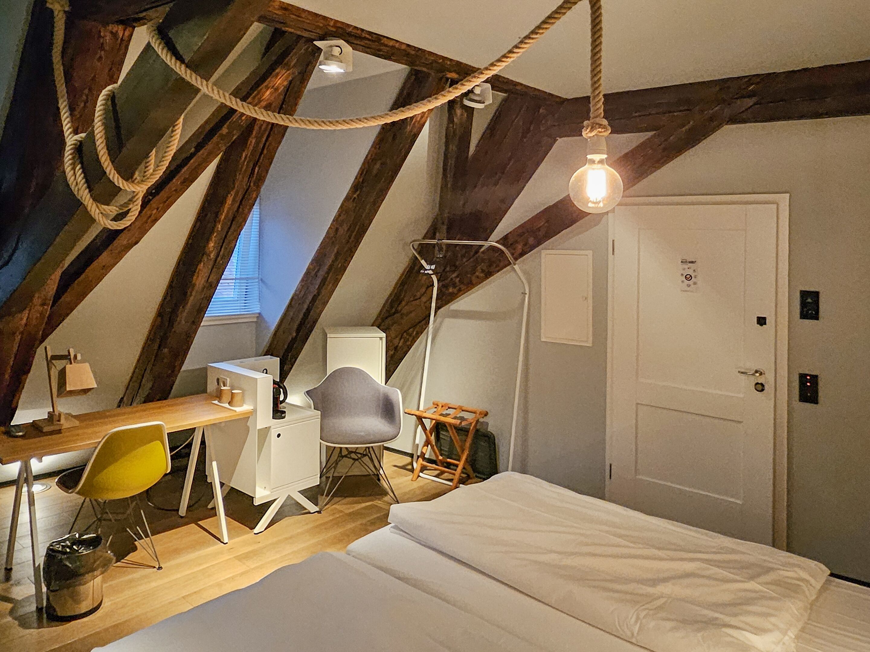 A loft-style bedroom with a sloped ceiling, exposed wooden beams, a desk with a chair, a bed, and a floor lamp.