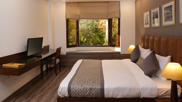 A hotel room with a bed, desk, chair, and a large window with a view of trees. - Ahuja Residency, Dlf Phase II (Gurugram)