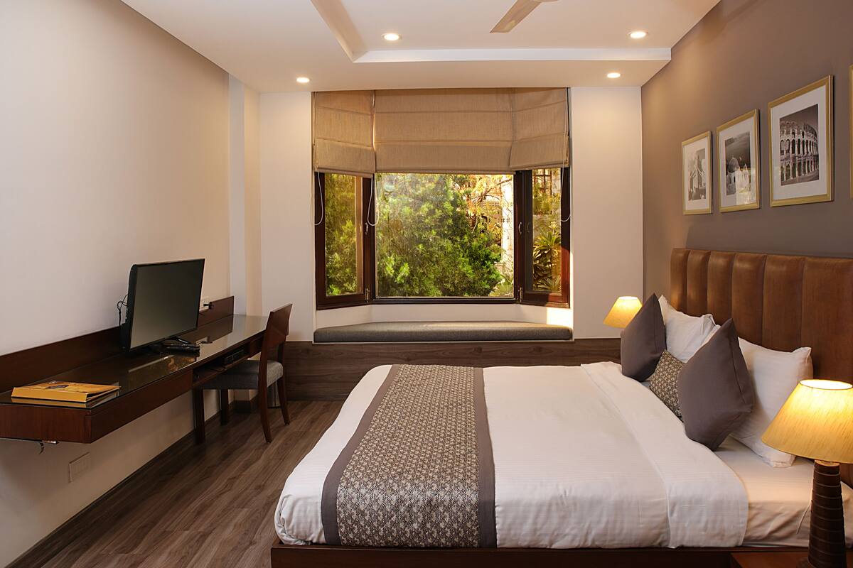 Habitación de hotel con cama, escritorio, silla y un amplio ventanal con vista a árboles.