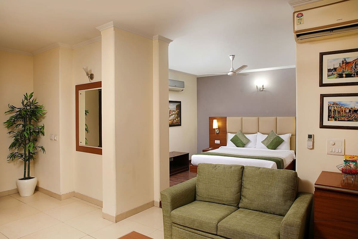 Habitación de hotel con cama, un sillón verde, una planta en maceta, ventilador de techo y aire acondicionado de pared.