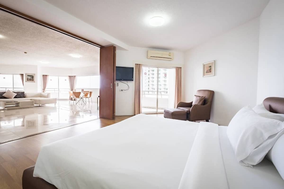 A modern hotel room with a large bed, a seating area with a sofa and chairs, and a view of the city through large windows.