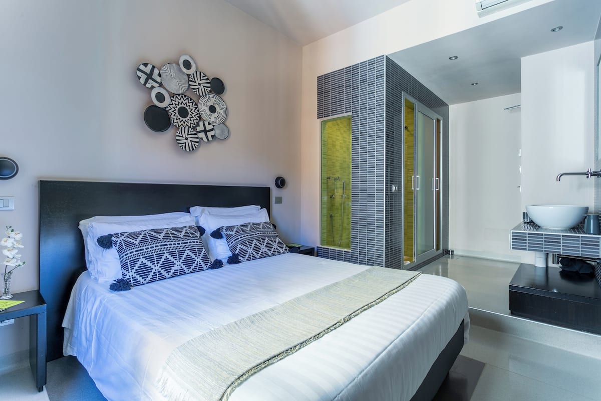 A modern bedroom with a large bed, a wall-mounted decorative piece, and a bathroom with a large mirror and sink.