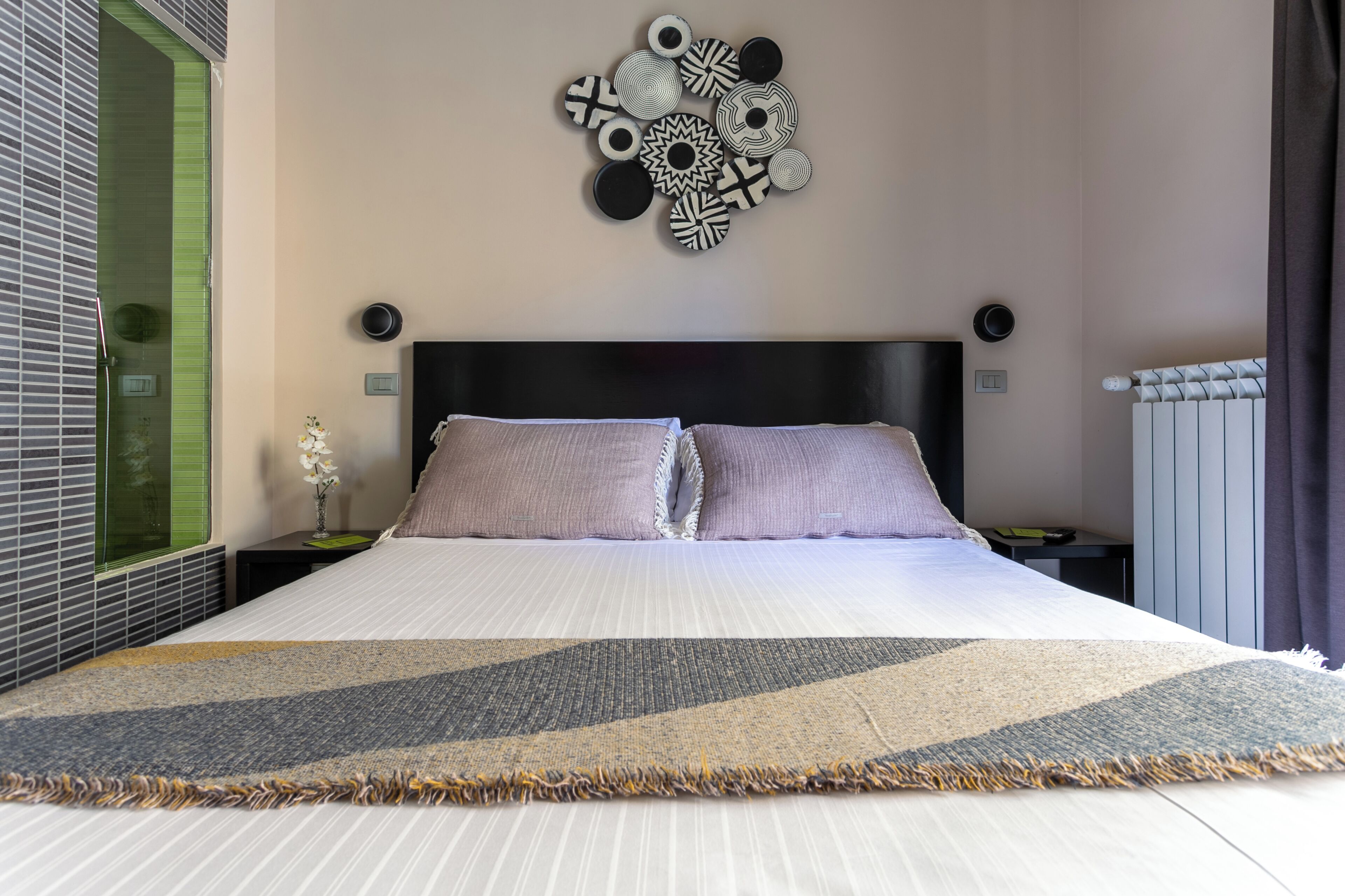 A neatly made bed with a patterned bedspread, a wall-mounted abstract art piece, and a bedside table with a lamp.