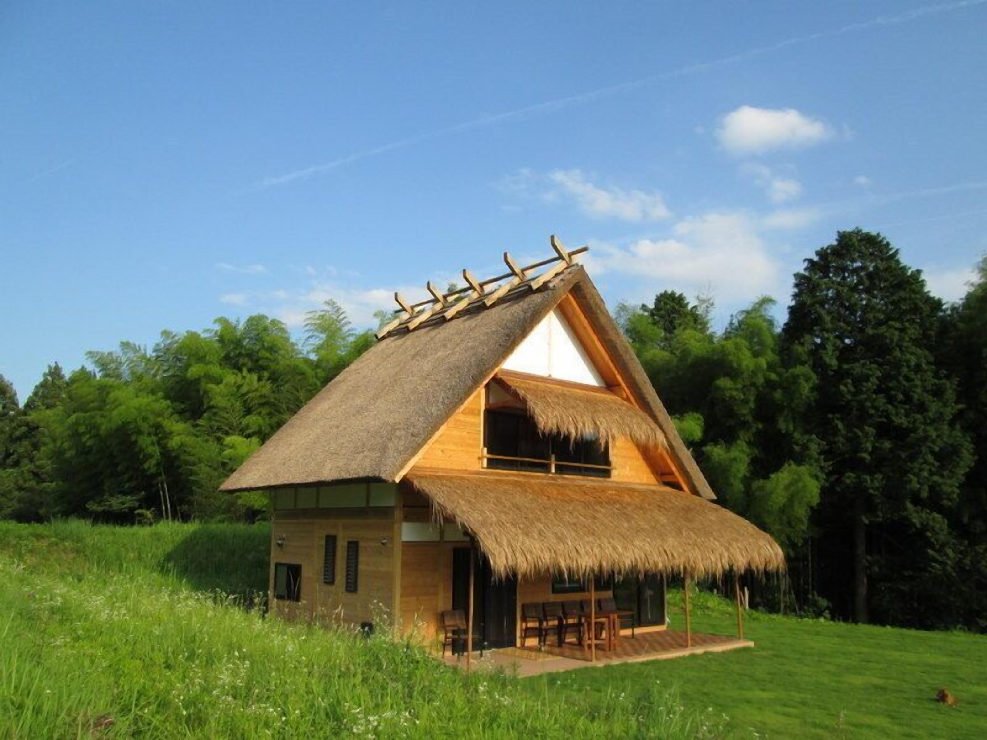 A thatched-roof house with a wooden exterior, a balcony, and a grassy field in the background.