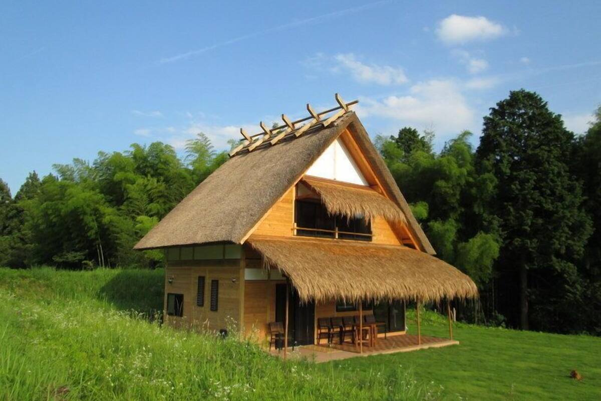 A thatched-roof house with a wooden exterior, a balcony, and a grassy field in the background.