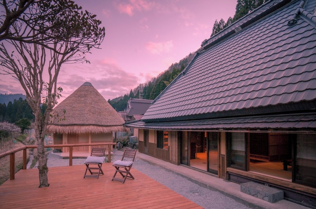 A wooden deck with two chairs, a thatched-roof structure, and a modern building with large windows.