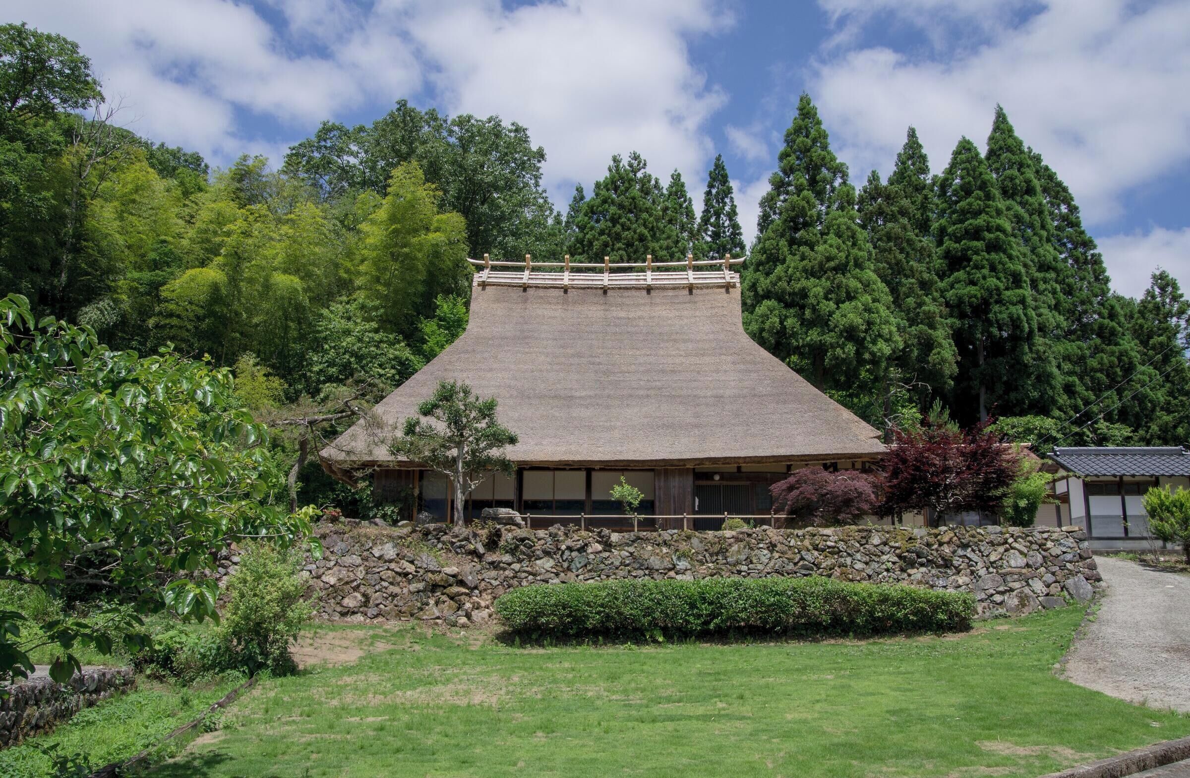 A traditional Japanese building with a sloped roof, surrounded by lush greenery and a stone wall.
