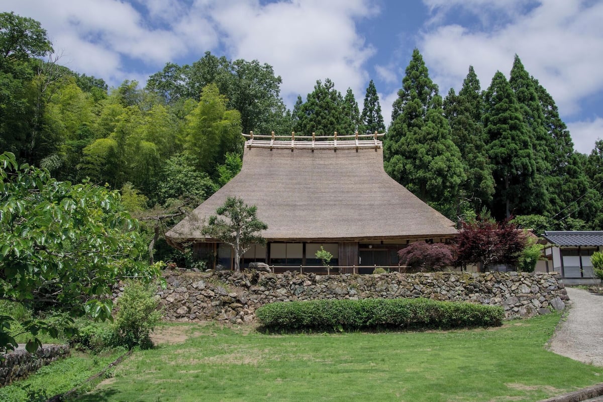A traditional Japanese building with a sloped roof, surrounded by lush greenery and a stone wall.