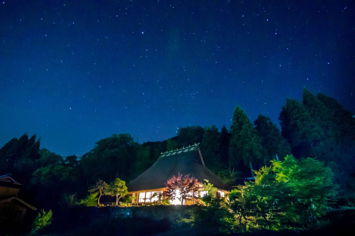 A traditional Japanese building with a thatched roof, surrounded by trees and a night sky with visible stars.