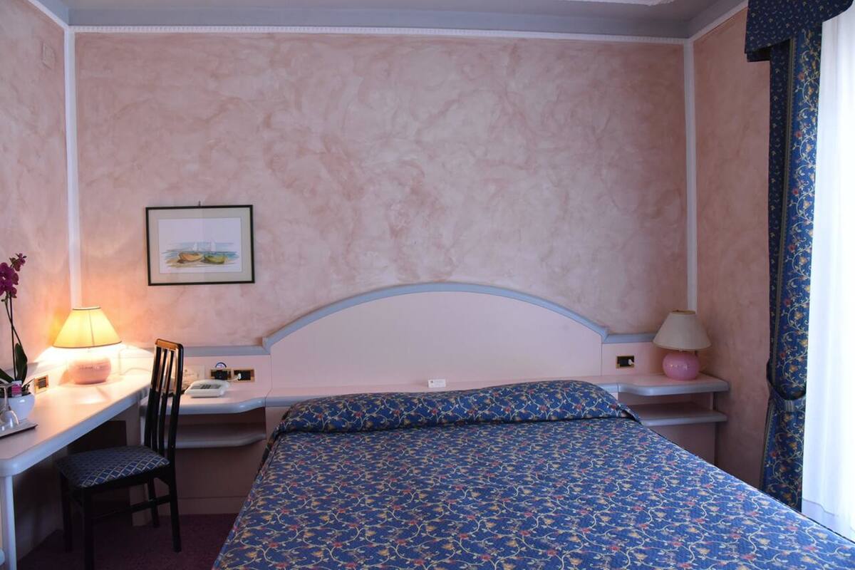 A hotel room with a bed, a desk, a chair, and a nightstand with lamps.