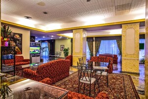 Un vestíbulo de hotel con sofás rojos de estampado, un comedor con mesa y sillas, y un televisor al fondo.