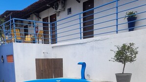 Une piscine avec un bord bleu, un banc blanc, un arbre en pot et un bâtiment doté d’une balustrade bleue et d’une porte en bois.