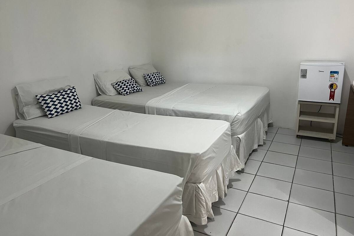 Ett hotellrum med tre enkelsängar, var och en med ett vitt överkast och en svartvit mönstrad kudde. Det står ett litet vitt kylskåp på ett träställ mot väggen.
