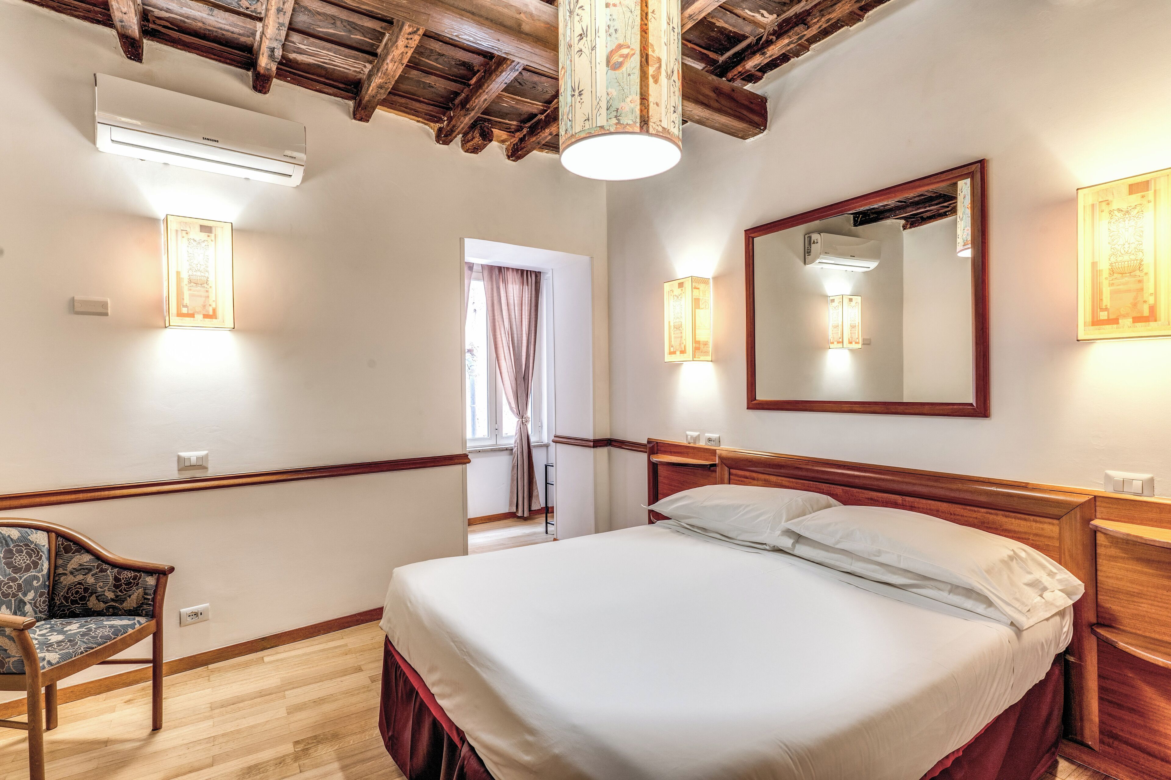 A hotel room with a large bed, wooden headboard, a wooden bench, a mirror, and wall-mounted lights.