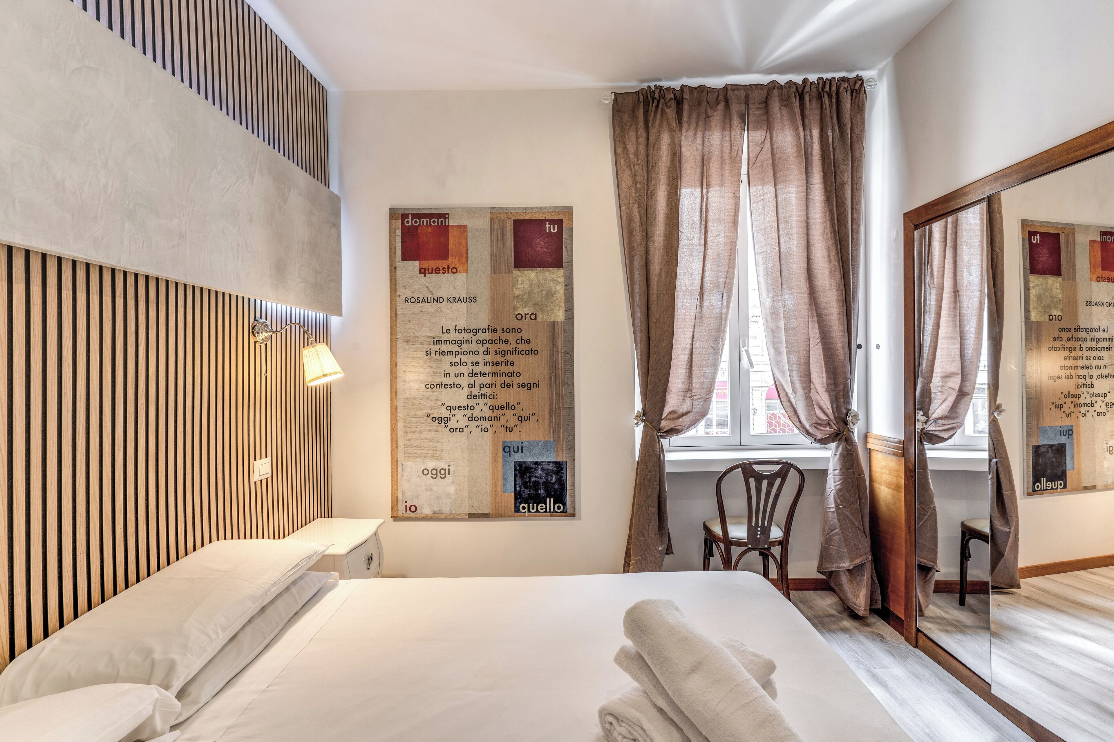 A hotel room with a bed, a wooden slat headboard, a wall-mounted artwork, a window with curtains, and a mirror.
