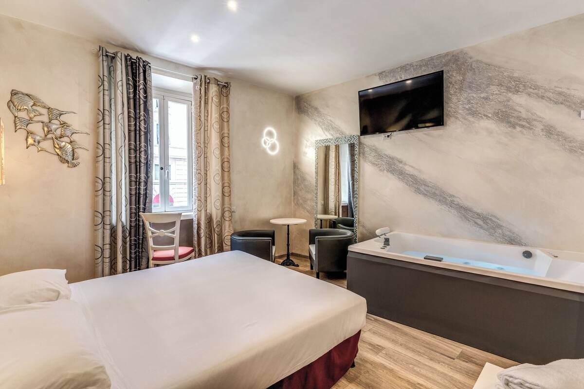 A hotel room with a bed, a sitting area with a table and chairs, a large bathtub, and a TV mounted on the wall.
