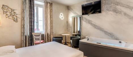Une chambre d’hôtel comprenant un lit, un coin salon avec une table et des chaises, une grande baignoire et un téléviseur fixé au mur.