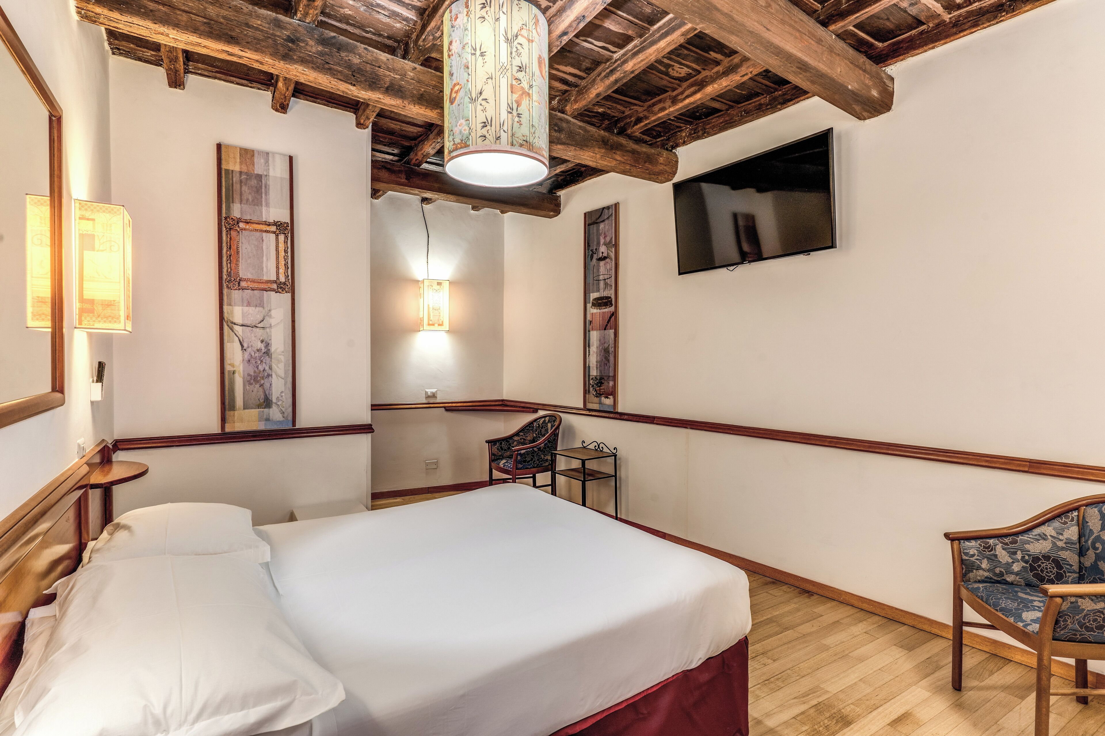 Une chambre d’hôtel avec un plafond en bois, un lit avec du linge de maison blanc, une petite table, une chaise et des œuvres d’art accrochées aux murs.