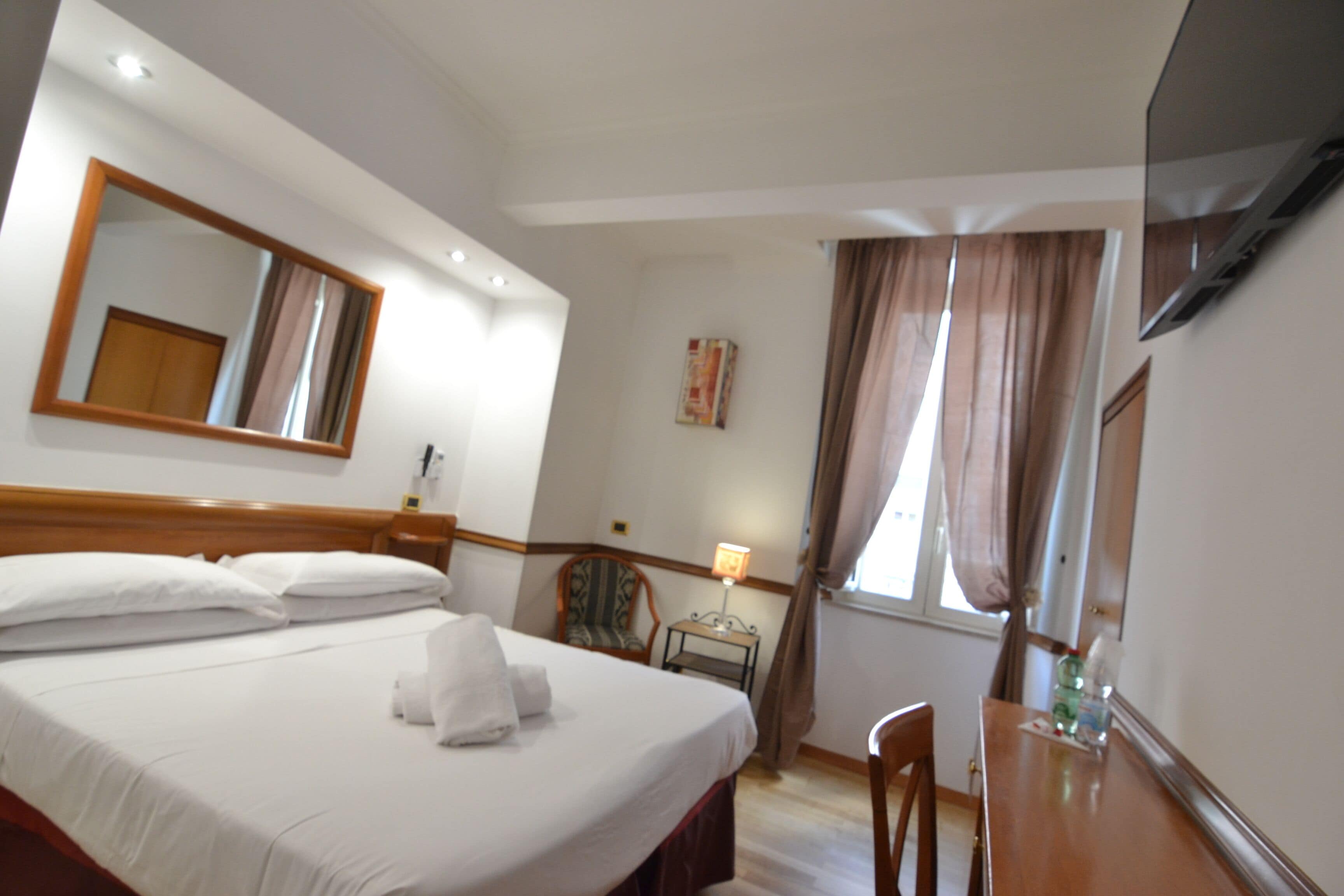 A hotel room with a bed, a desk, a chair, a mirror, a TV, and a window with curtains.