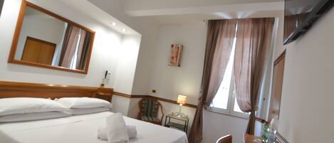 A hotel room with a bed, a desk, a chair, a mirror, a TV, and a window with curtains.