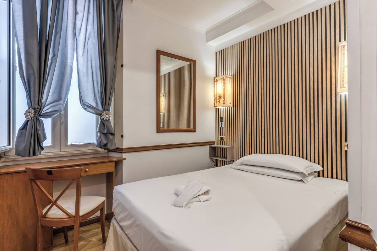 A hotel room with a bed, a wooden desk, a chair, a mirror, and a wall-mounted light fixture.