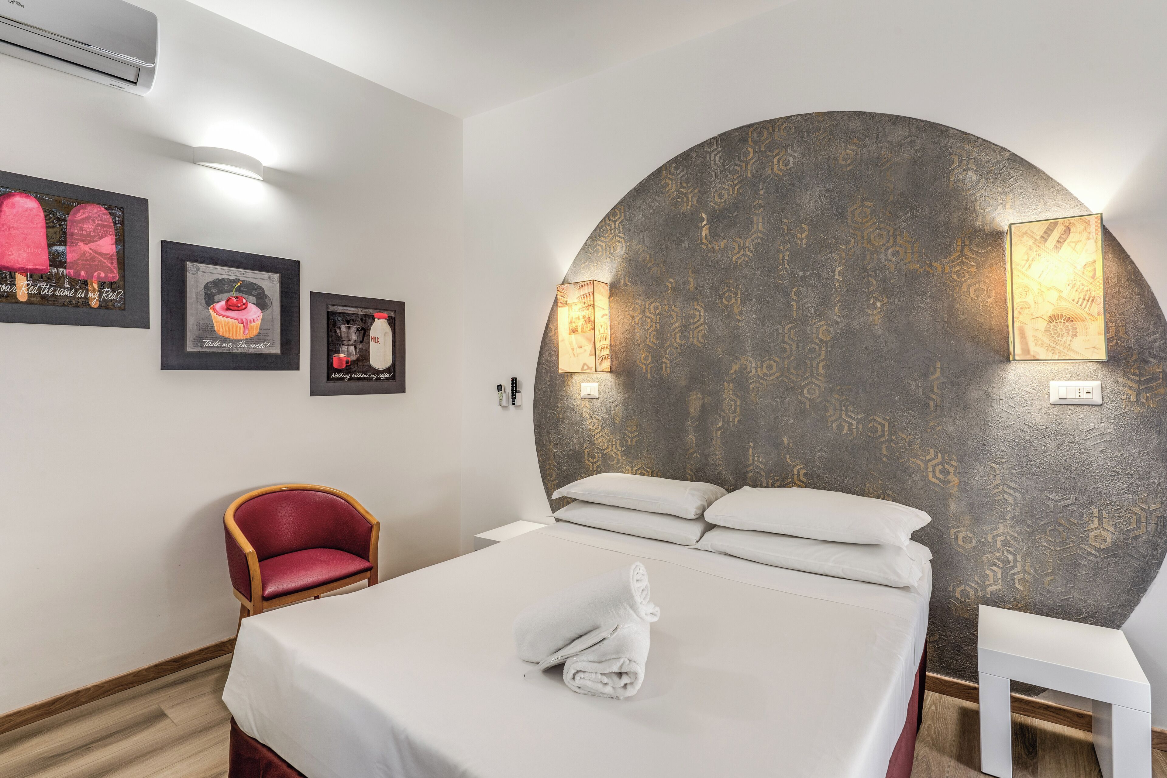 A hotel room with a bed, a red chair, a white table, and wall-mounted artwork.