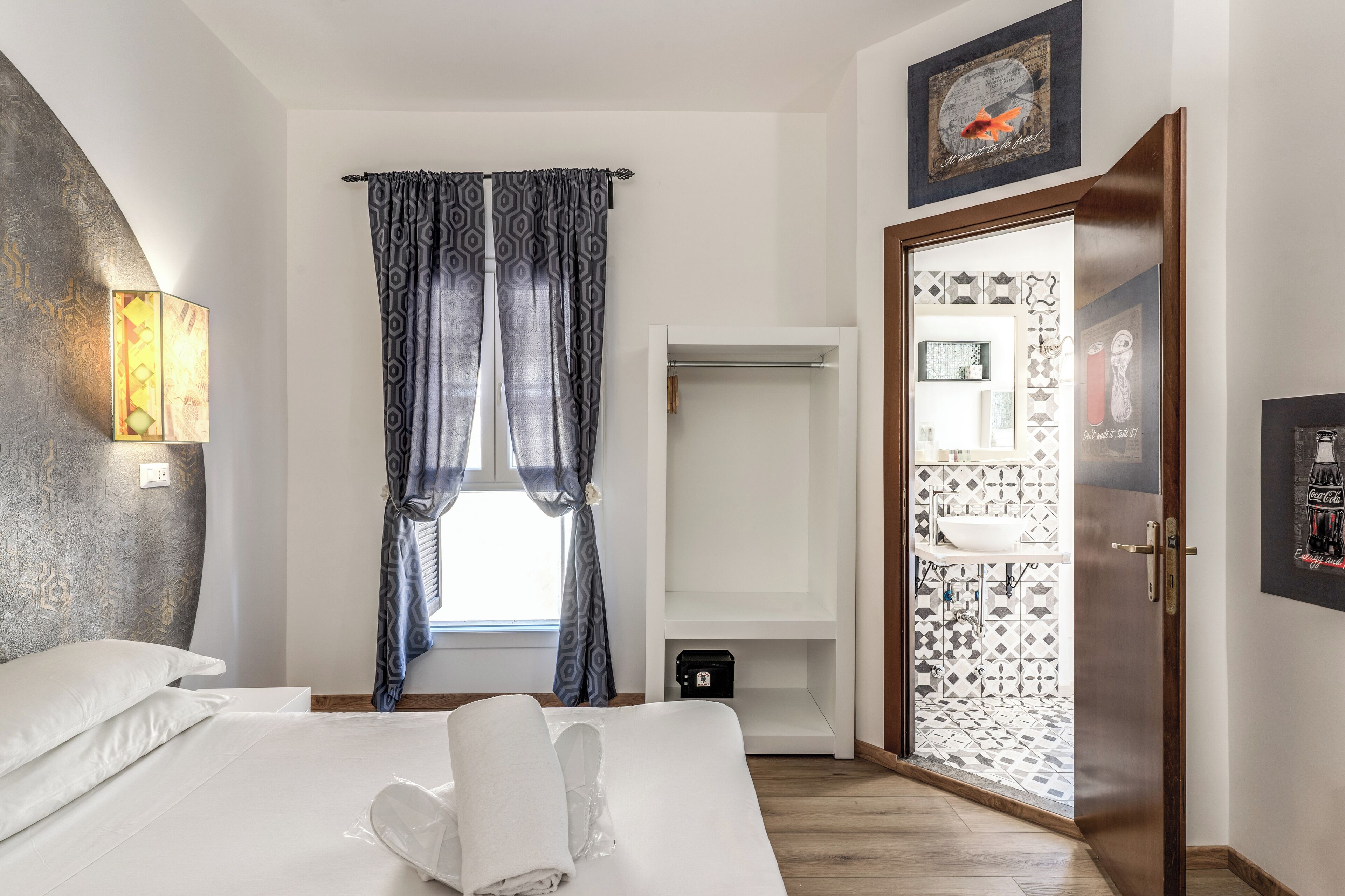 A hotel room with a bed, a window with patterned curtains, a wardrobe, and a bathroom visible through an open door.