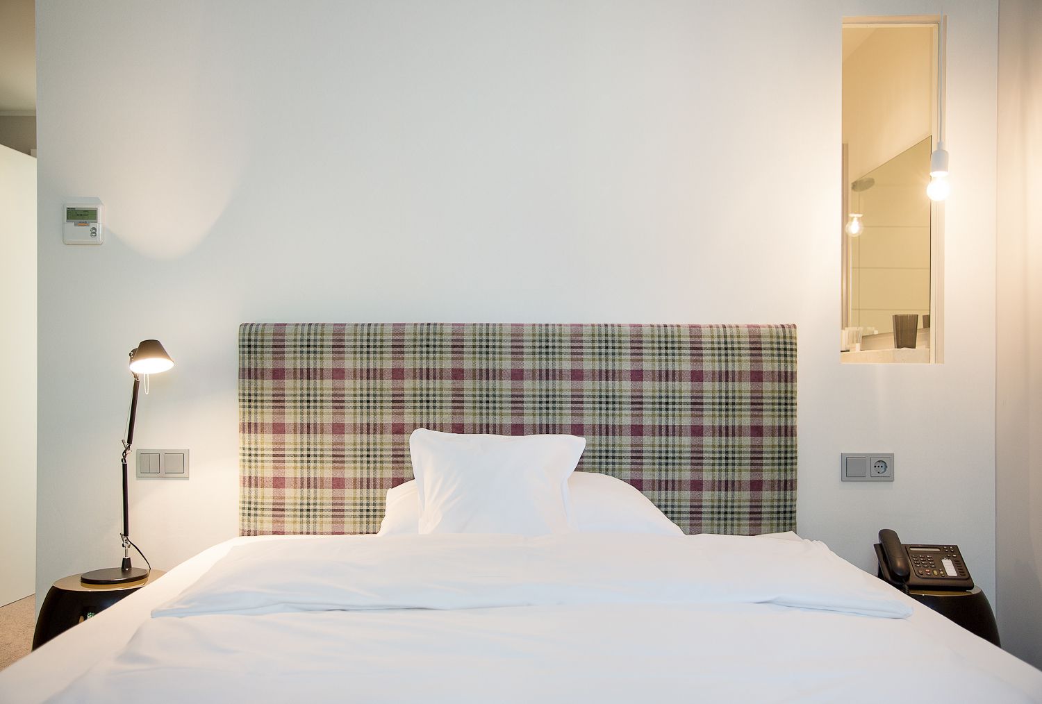 A neatly made bed with a plaid headboard, a bedside lamp, a phone on a nightstand, and a mirror on the wall.
