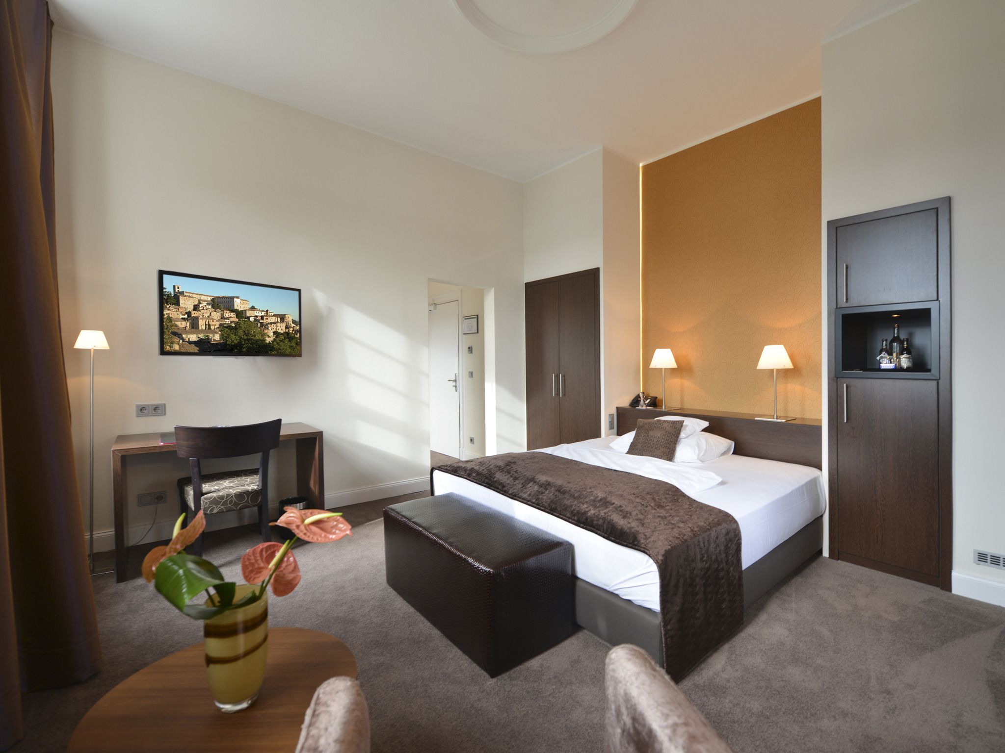 Een hotelkamer met een bed, nachtkastjes, een bureau, een stoel, een bijzettafeltje met een vaas en een aan de muur hangend schilderij.