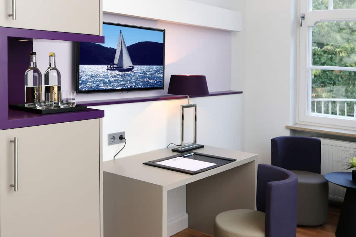 A modern room with a desk, a television displaying a sailboat on water, a lamp, and a window with a view of greenery.