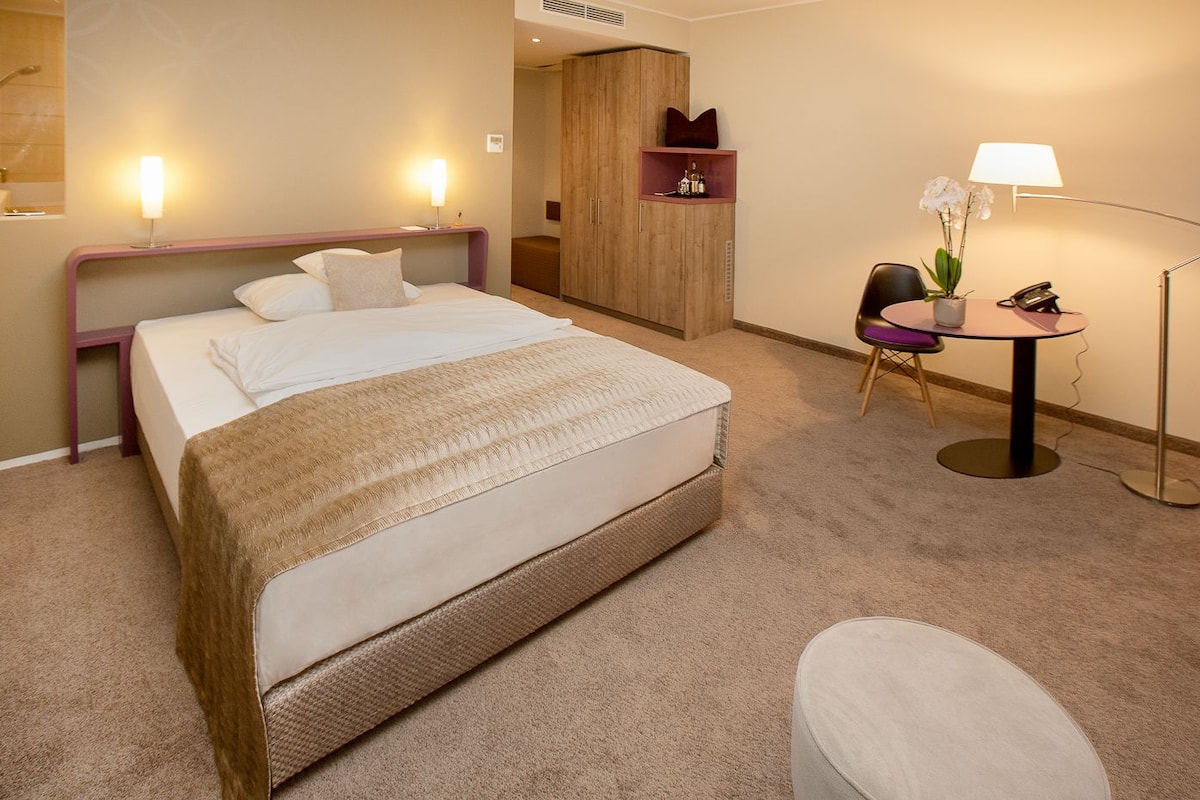 A hotel room with a large bed, bedside lamps, a small table with a lamp, a chair, and a wardrobe.