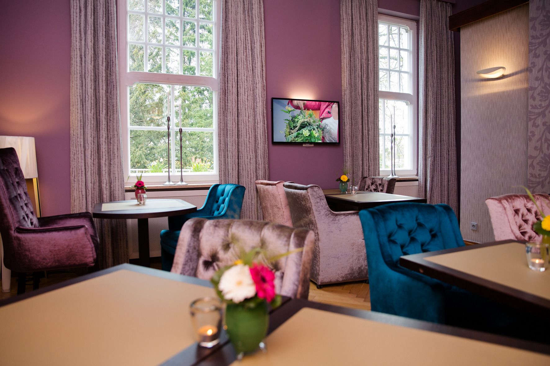 A room with a dining area, a TV mounted on the wall, and large windows with curtains.