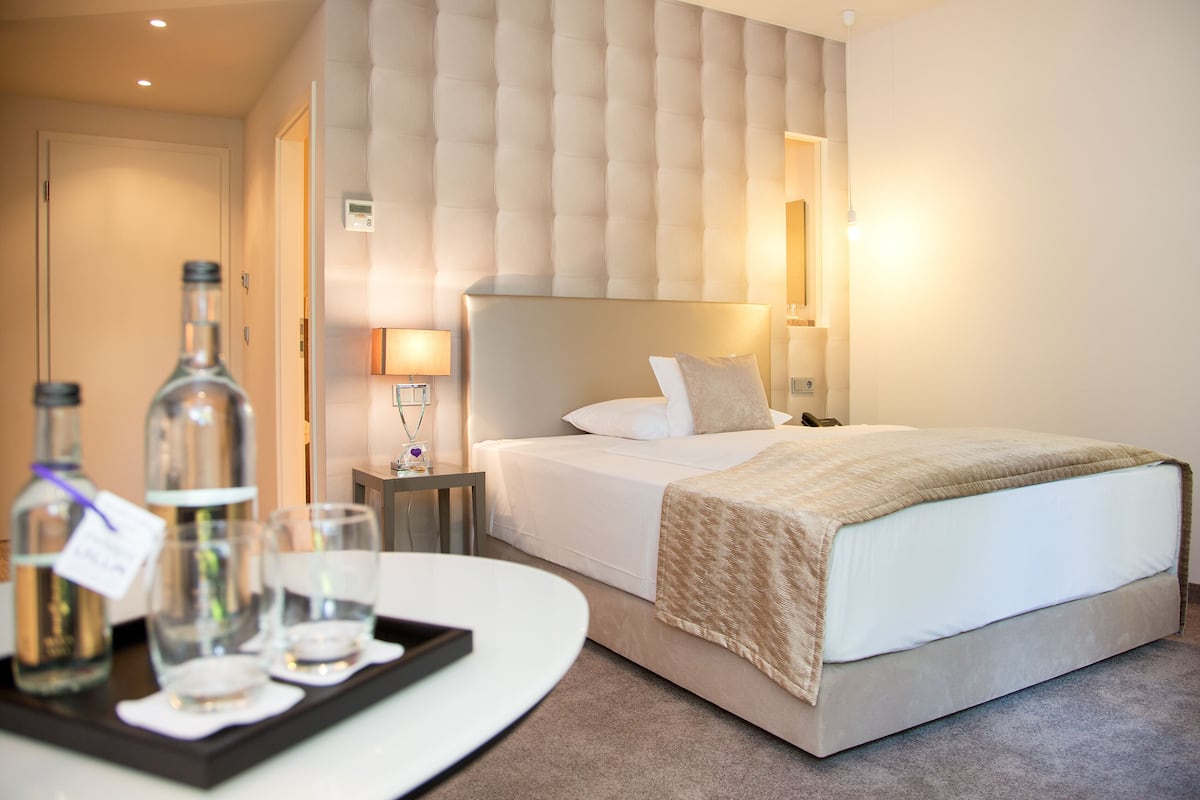 A hotel room with a large bed, a bedside table with a lamp, a tray with bottles and glasses, and a wall-mounted thermostat.