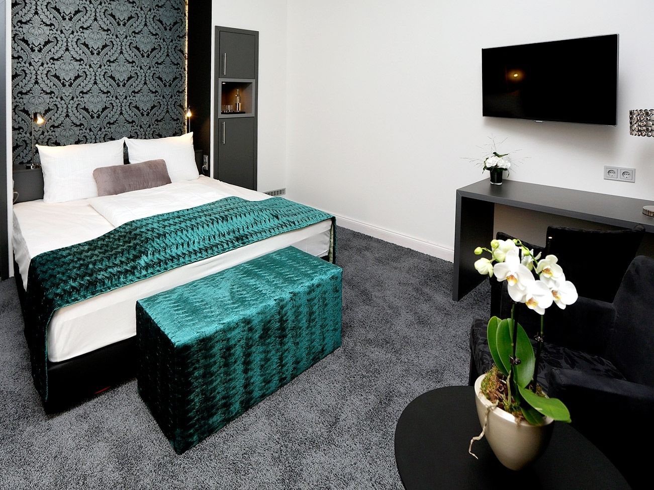 Habitación de hotel moderna con una cama bien hecha, una pequeña zona de estar, un televisor de pantalla plana y una orquídea en maceta.