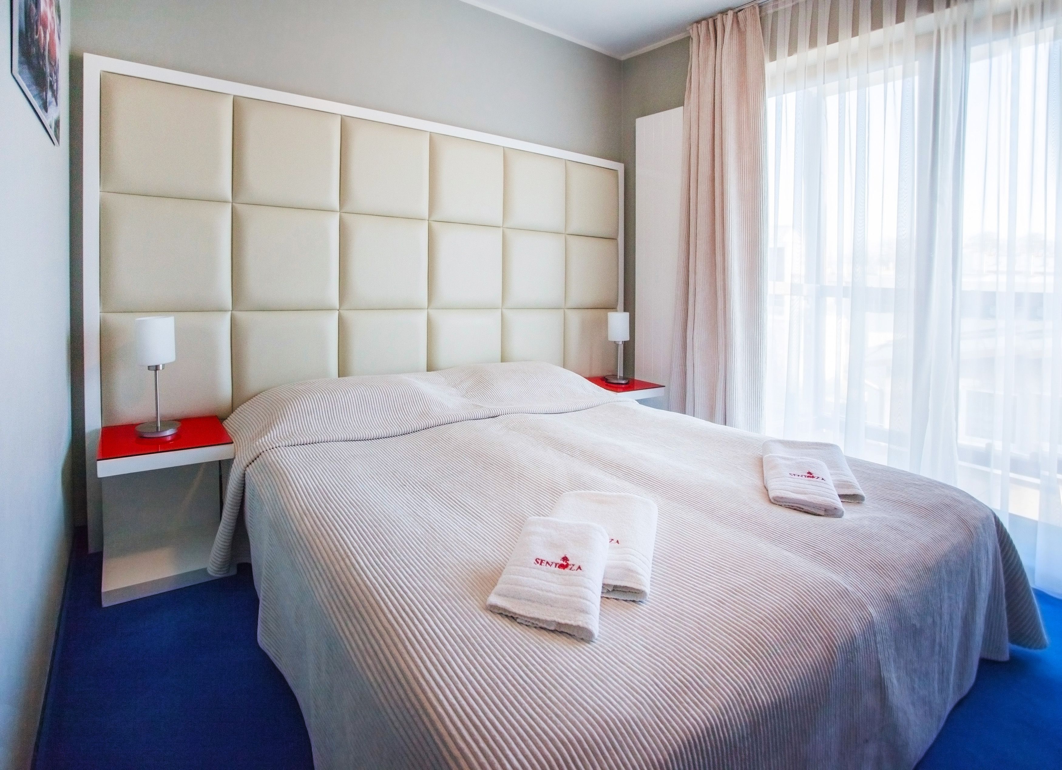 A hotel room with a large bed, two bedside tables, a headboard, and a window with sheer curtains.