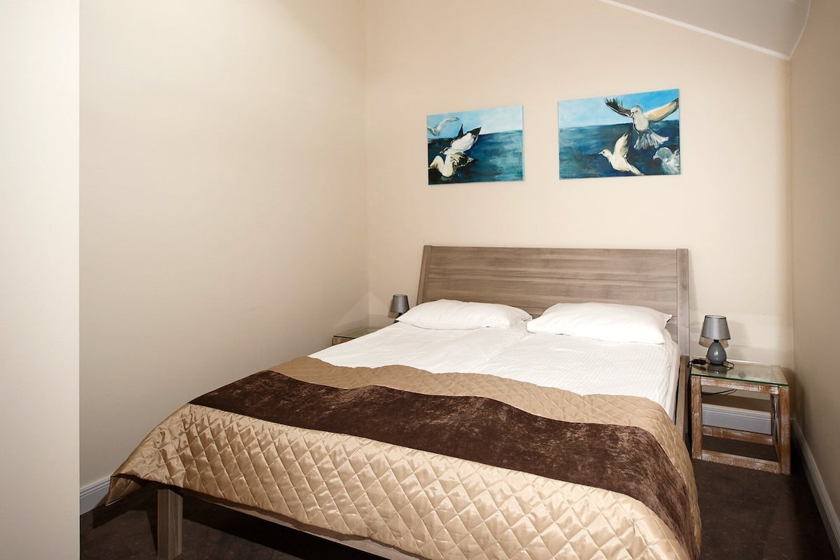 A neatly made bed with a quilted cover, a bedside table with a lamp, and two wall-mounted paintings of seagulls.