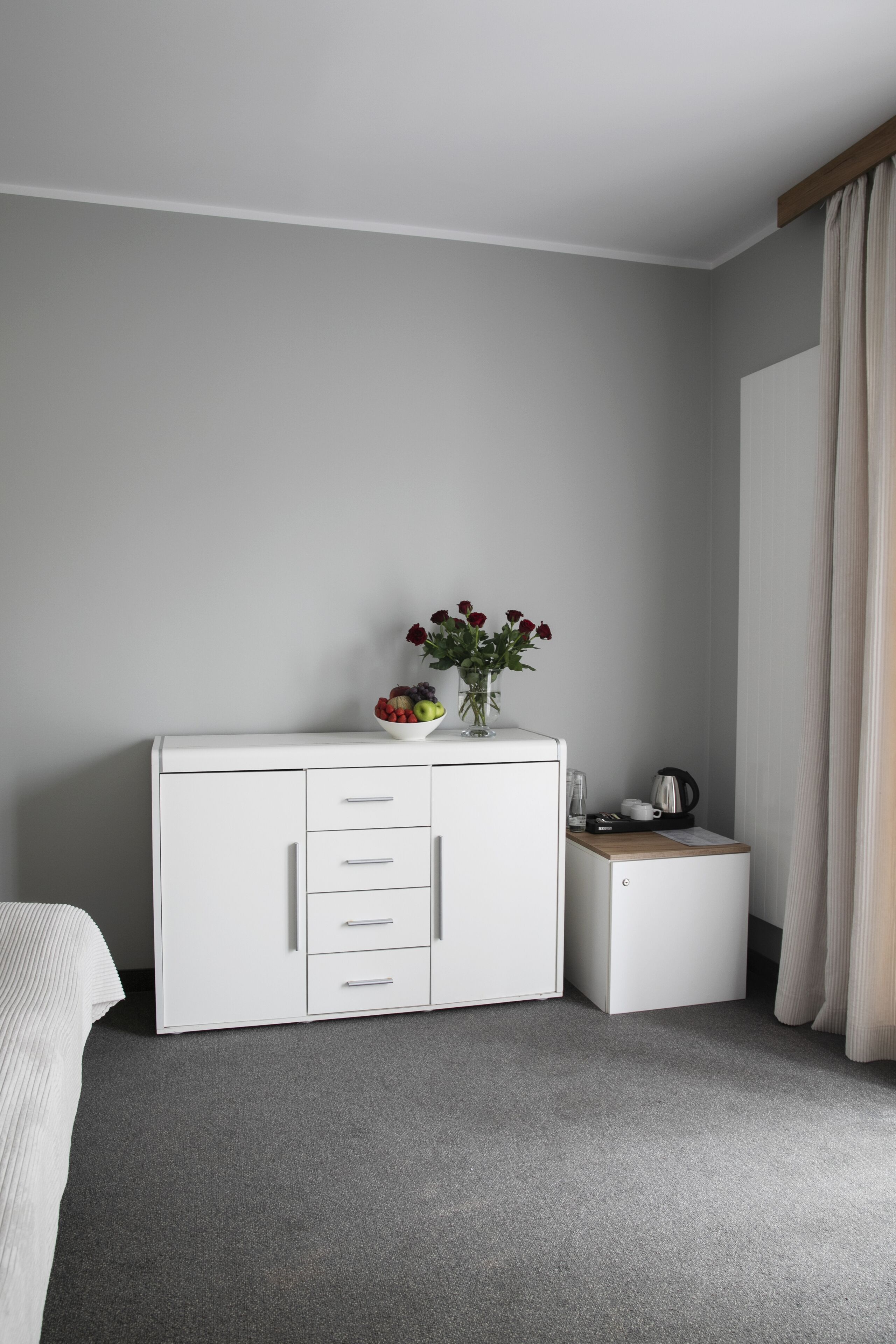 Un dormitorio con un armario blanco, una mesita de noche pequeña, una cama y una ventana con cortinas.