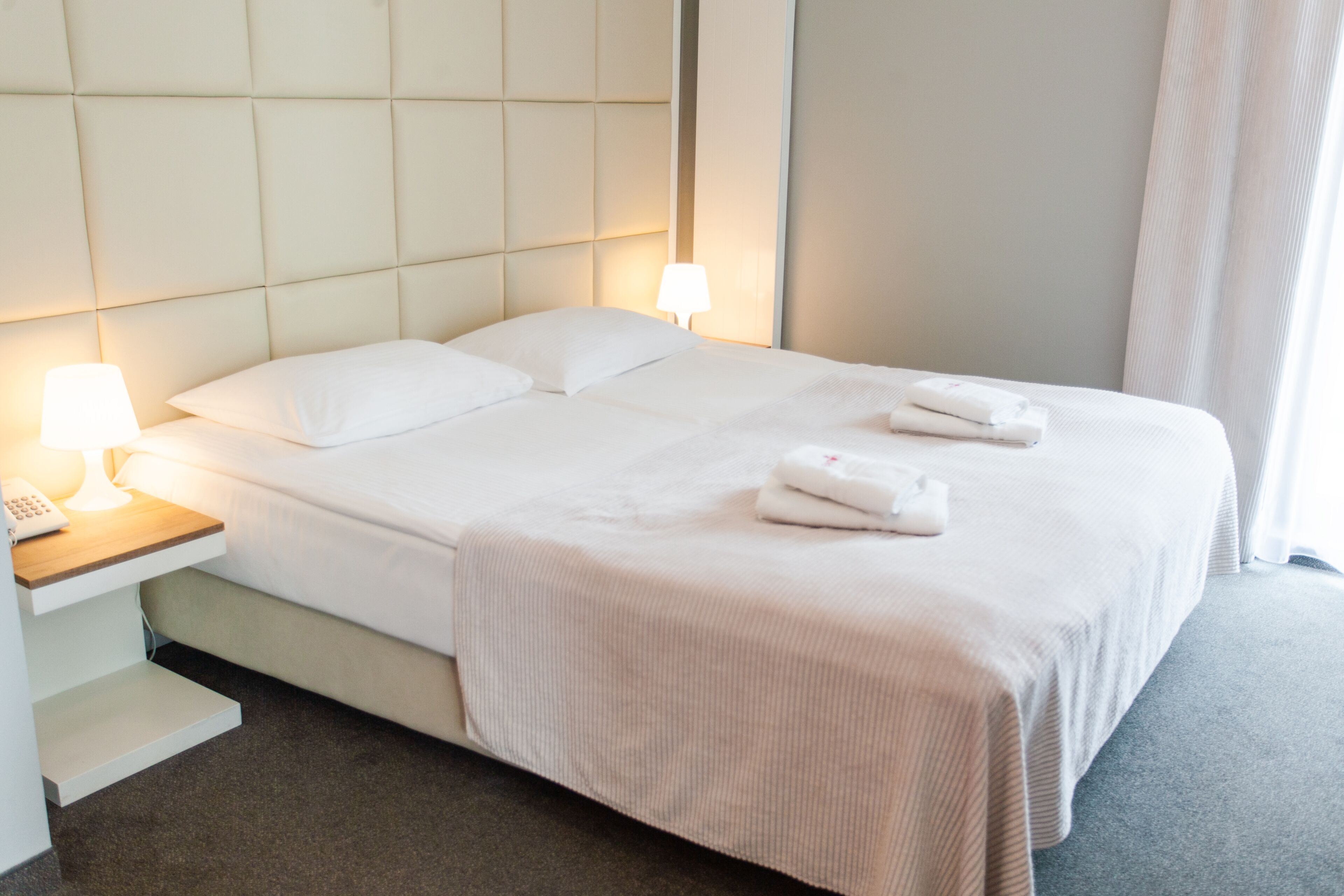 A neatly made bed with white linens, a bedside table with a lamp, and a folded towel on the bed.