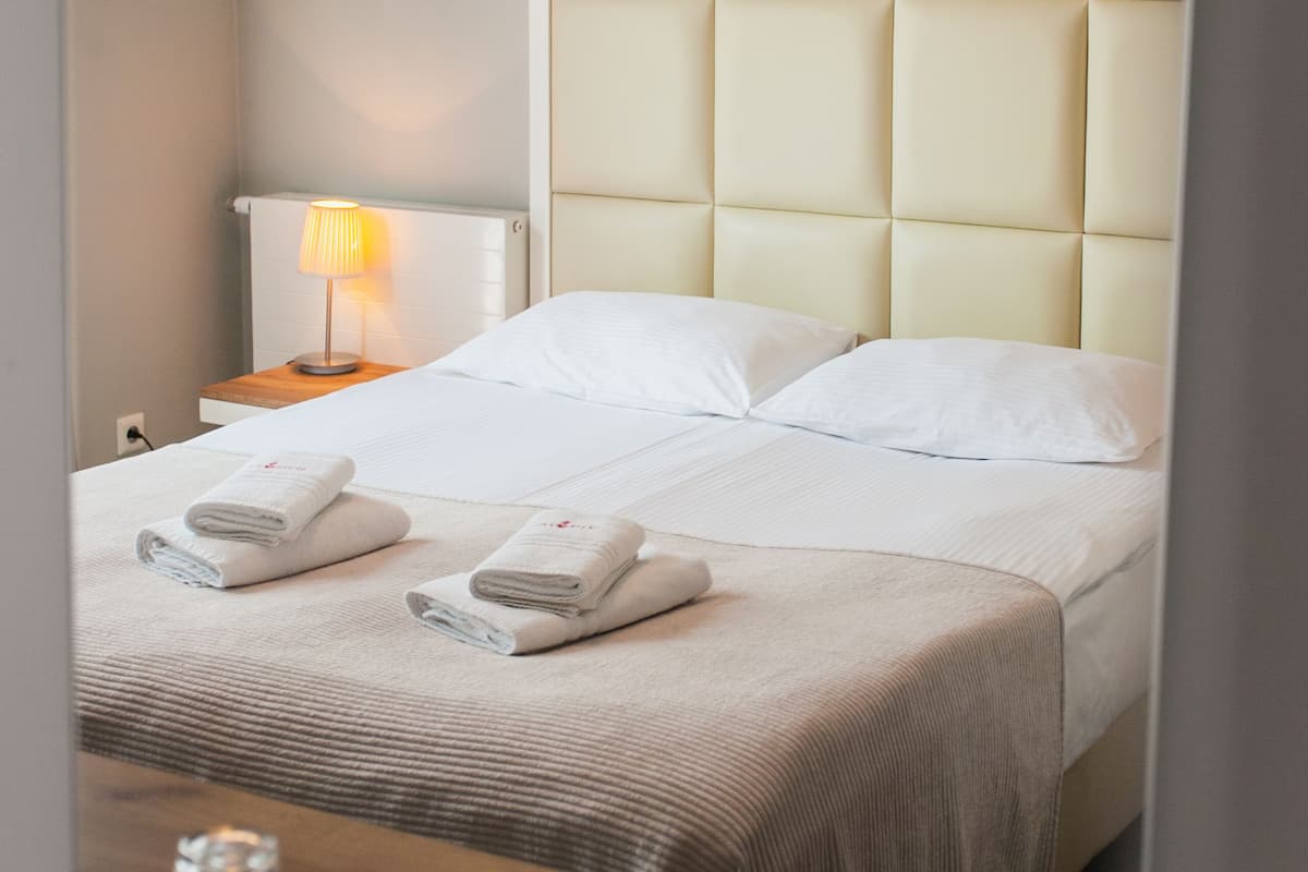 A neatly made bed with white linens and towels, a bedside table with a lamp, and a wooden nightstand.