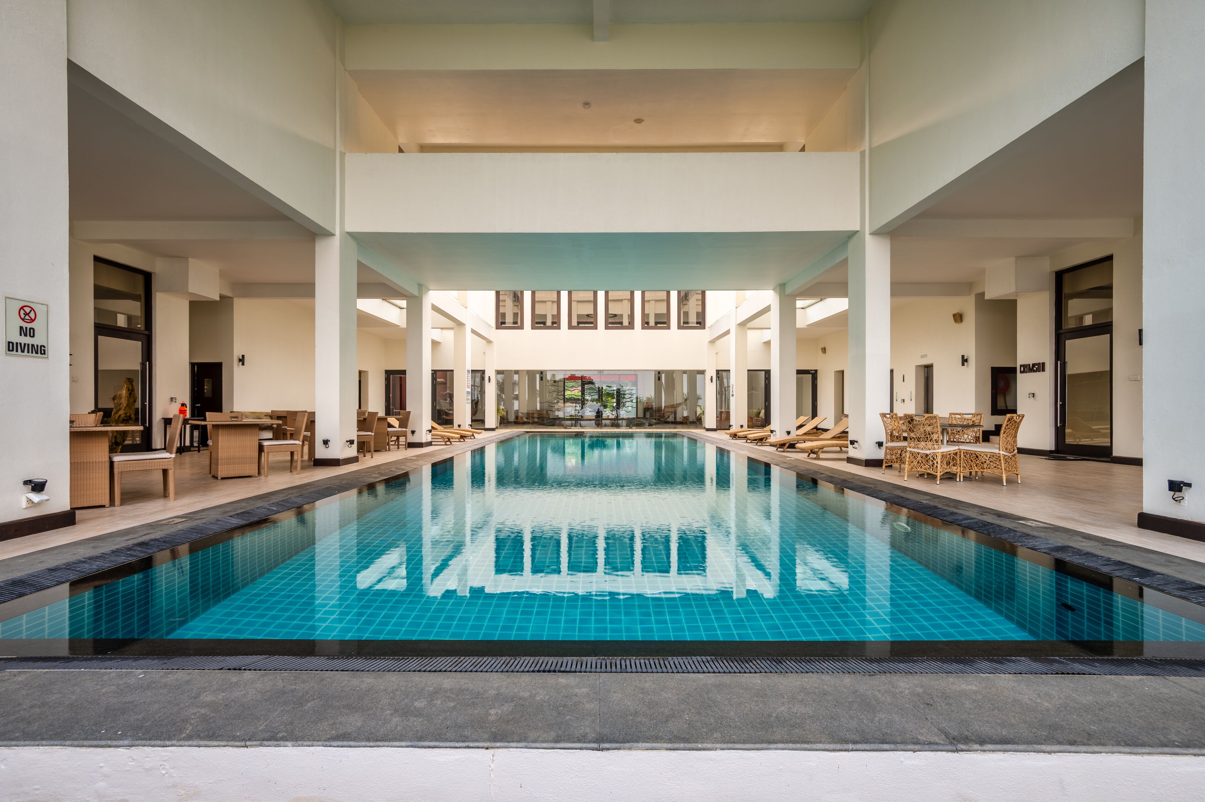 Une grande piscine intérieure, bordée de chaises longues et de tables de part et d’autre, et entourée de murs et de portes blancs.