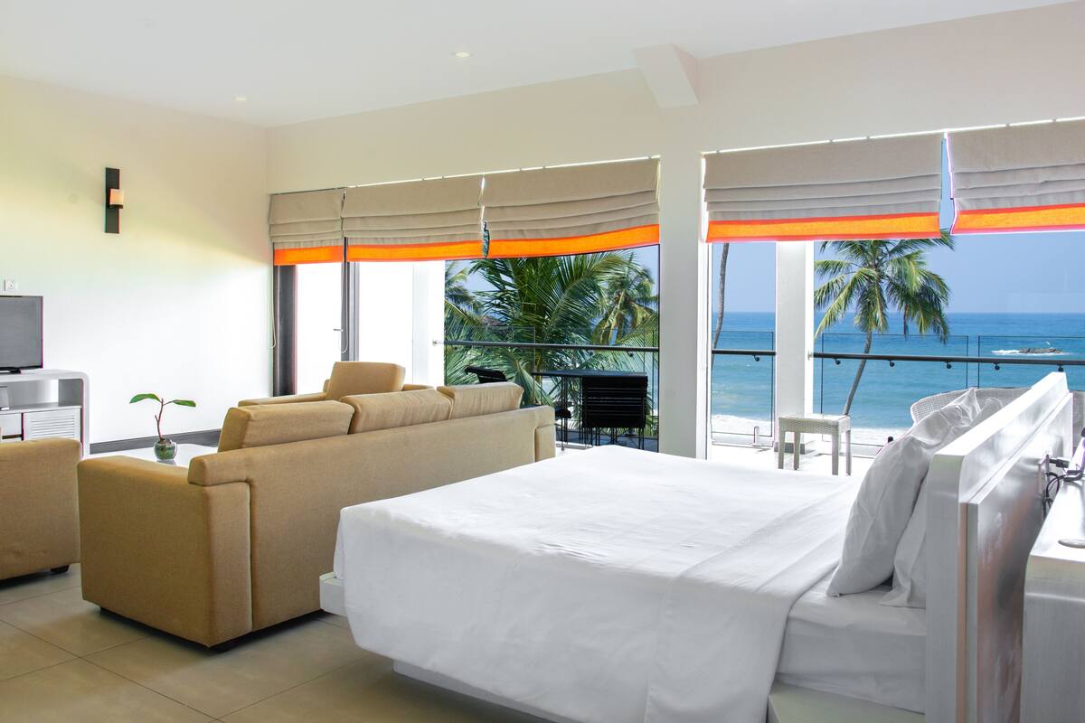 A hotel room with a bed, a sofa, a TV, and a balcony with a view of the ocean.