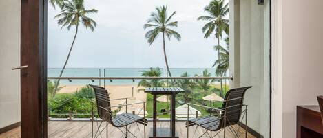 A balcony with a table and two chairs, overlooking a beach with palm trees and the ocean.