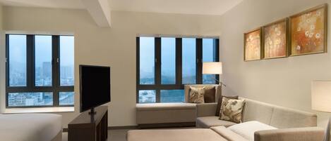 Habitación de hotel moderna con una cama grande, un sofá, un televisor y un escritorio. Amplias ventanas con vistas a la ciudad.