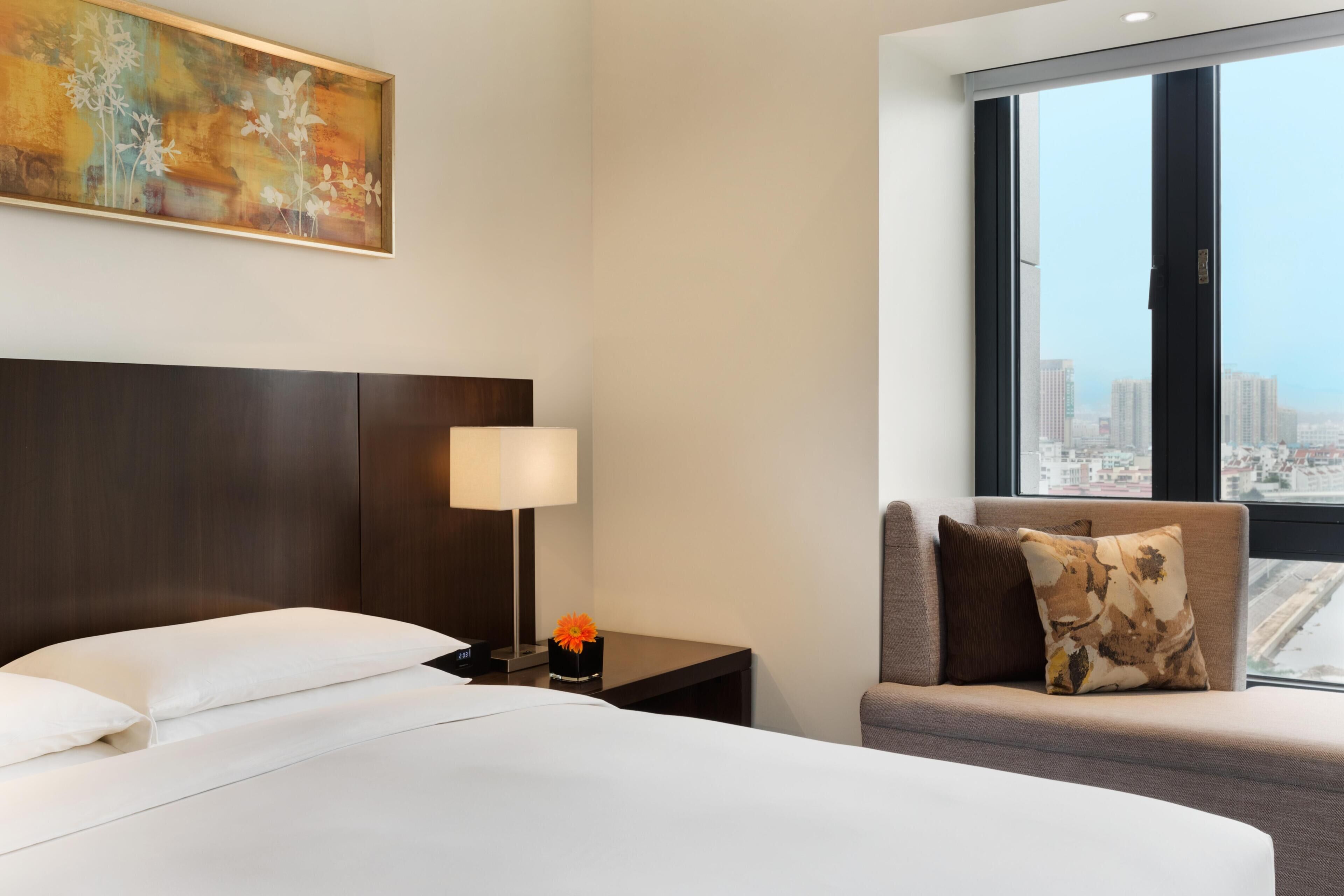 A hotel room with a large bed, a nightstand with a lamp, a small table with a flower, a sofa with cushions, and a city view through a large window.