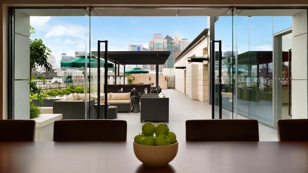 Ein modernes Hotelzimmer mit einem großen Fenster, einem Esstisch mit einer Schüssel Äpfel und Blick auf die Stadt sowie eine Außenbestuhlung.
