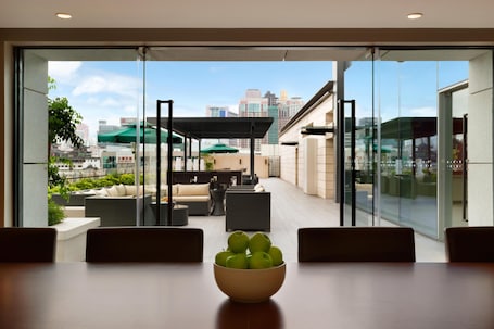 A modern hotel room with a large glass window, a dining table with a bowl of apples, and a view of the cityscape and outdoor seating area.