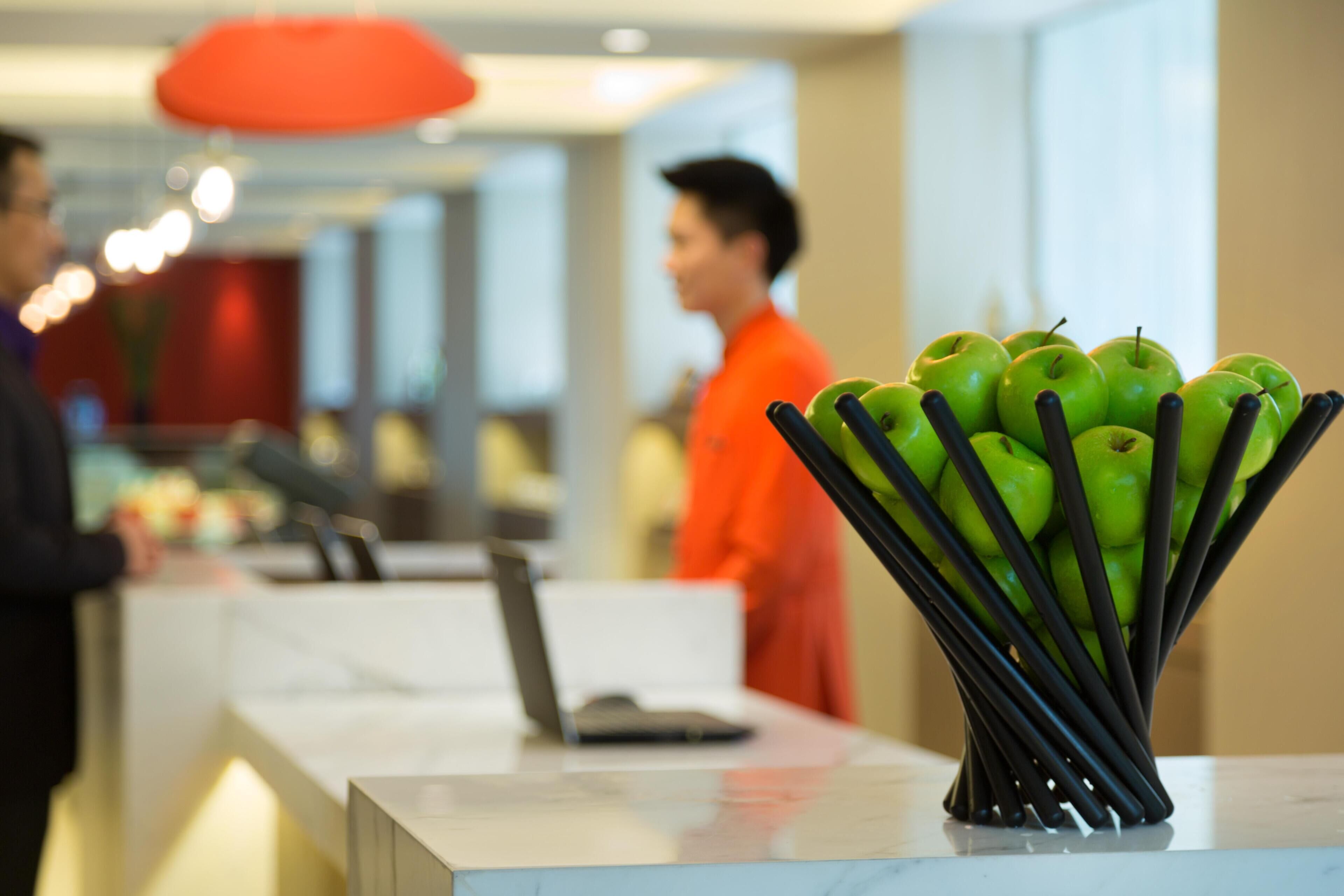 A reception desk with a laptop, a vase of green apples, and a person in an orange uniform.