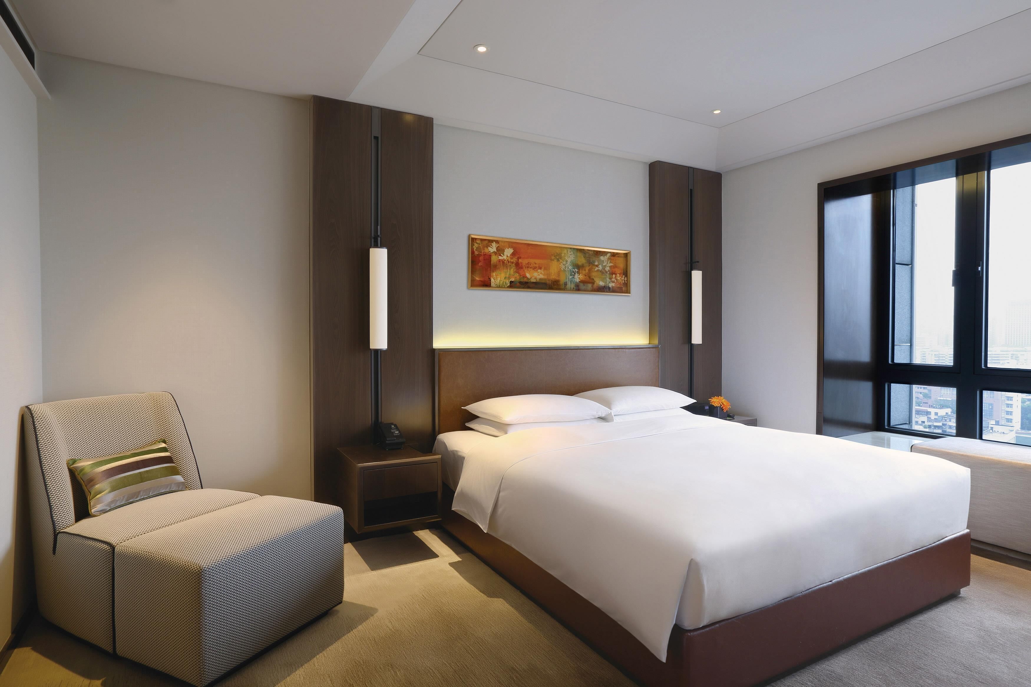 A hotel room with a bed, a chair, a nightstand, a lamp, a painting on the wall, and a large window.