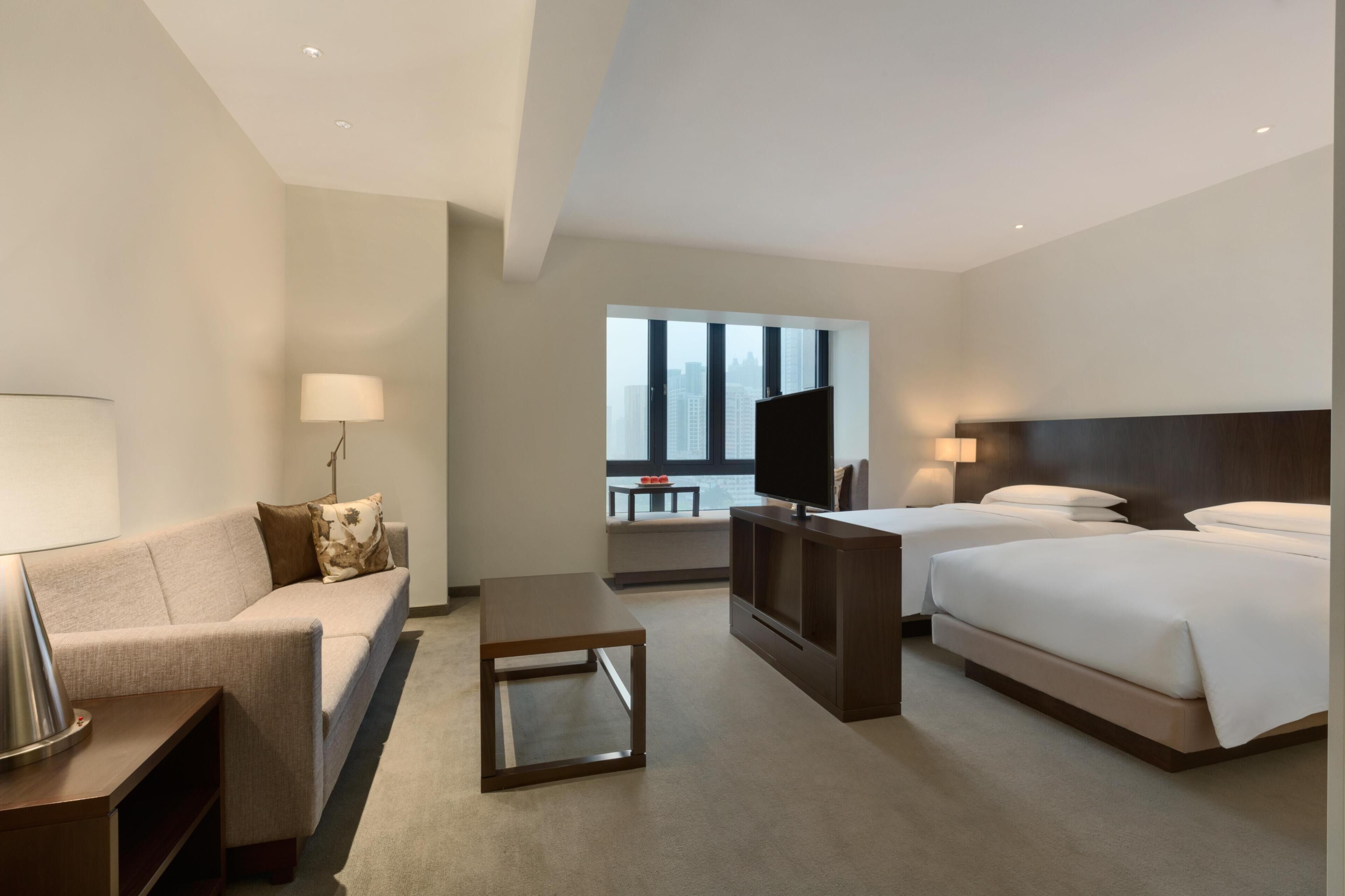 A hotel room with a sofa, a TV, a bed, and a window with a view of buildings.